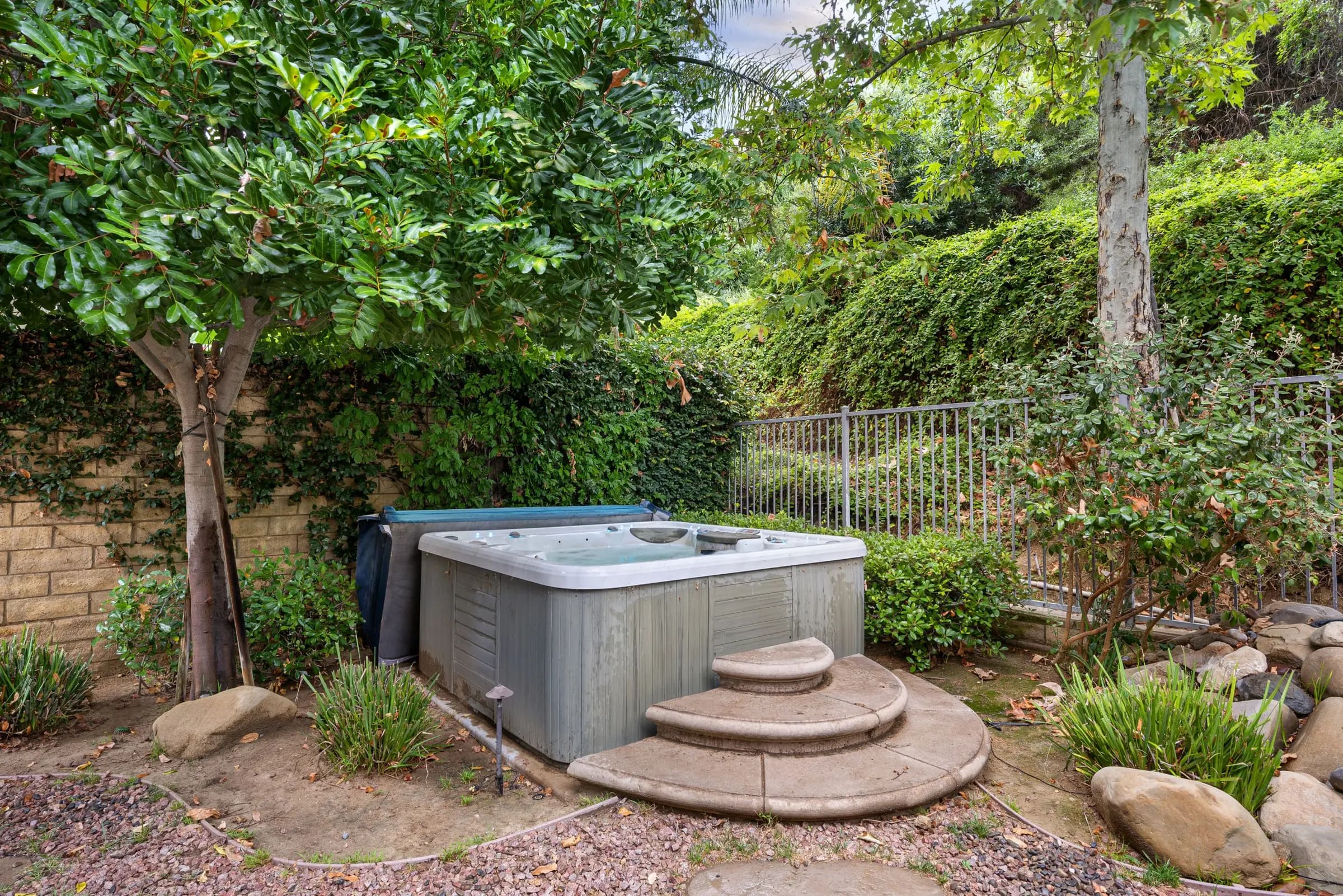 A weathered spa with its cover folded behind it, sitting beside a block wall and an iron fence in a leafy backyard