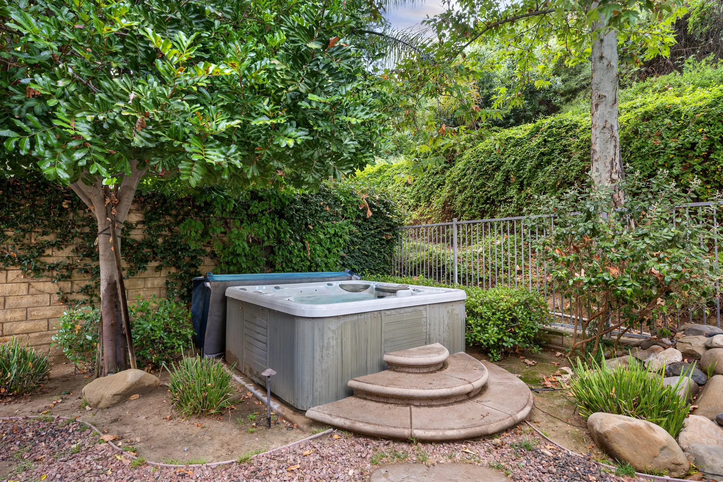 A weathered hot tub tucked against an iron fence, with curved stone steps leading up to it