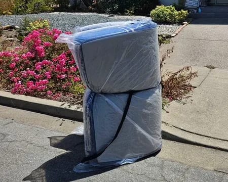 Two mattresses wrapped in plastic standing at the curb beside a flower bed, ready for pickup