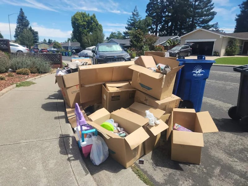 Moving boxes and loose household things stacked on a curb, with houses and parked cars across the street