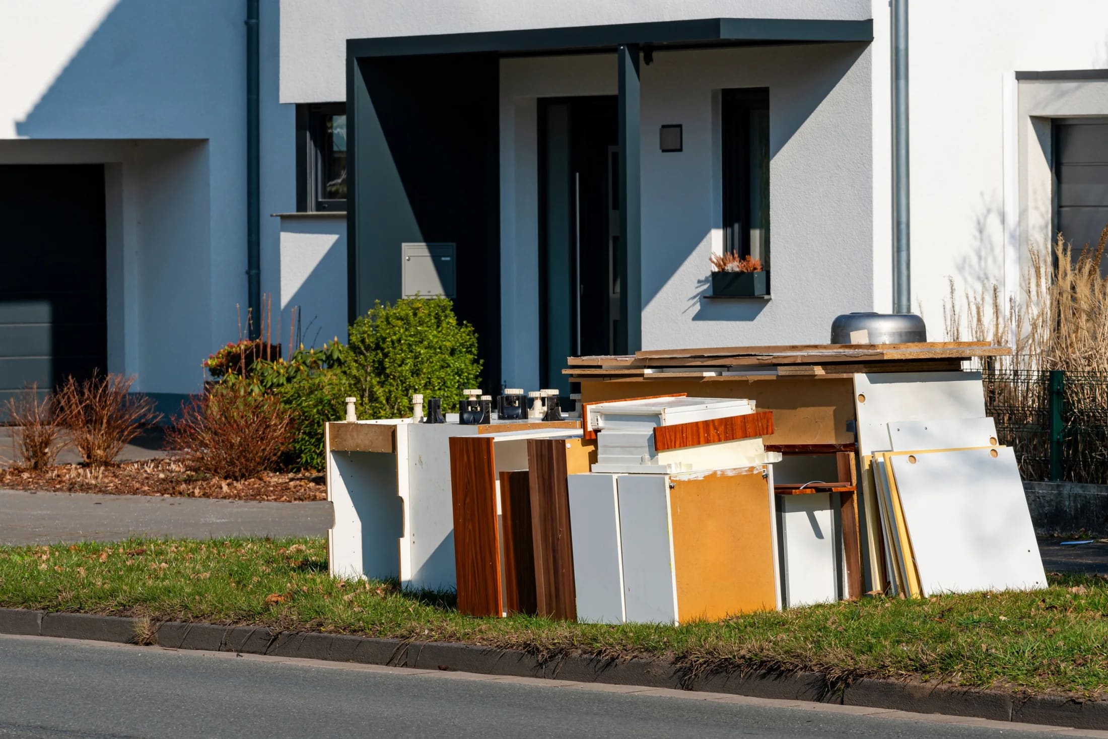 A pile of worn cabinet doors and particleboard shelves at the curb outside a house with a dark front entry