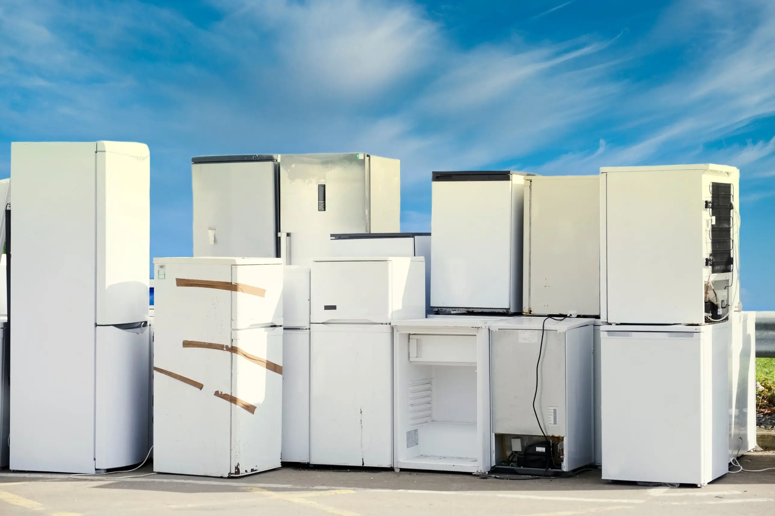 A row of old white refrigerators and freezers lined up outdoors under a bright blue sky