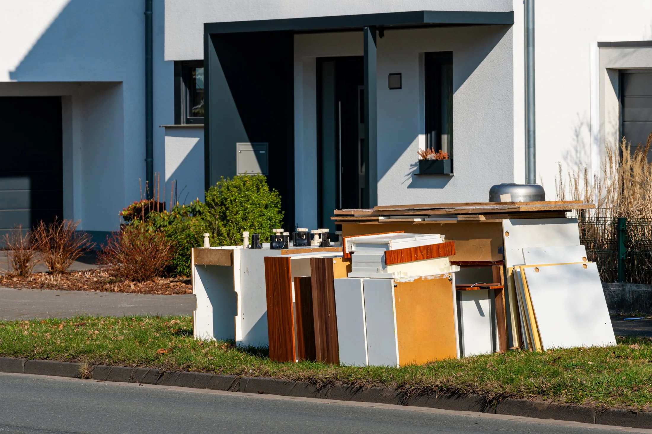 Old cabinets, drawers and furniture panels stacked on the grass in front of a modern white house on a sunny day