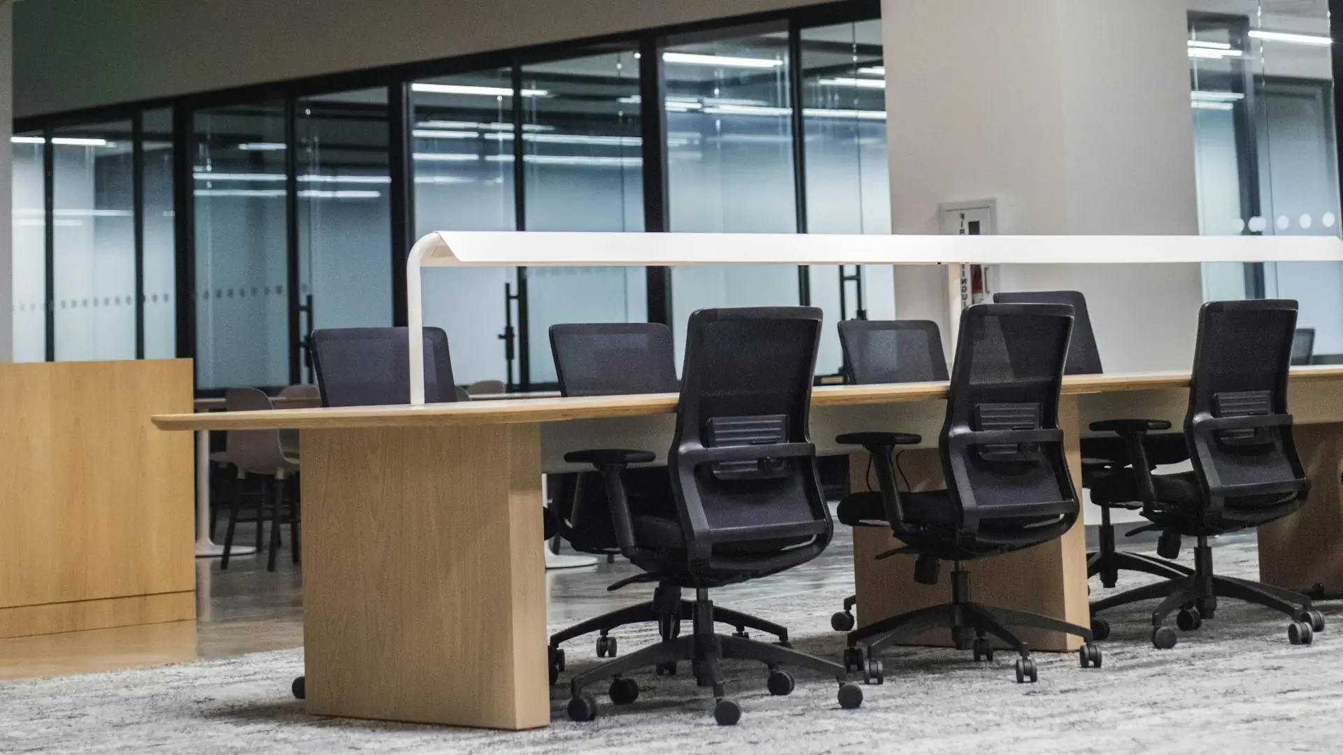 Rows of black mesh office chairs at a long shared desk in an open office, with glass-walled meeting rooms behind