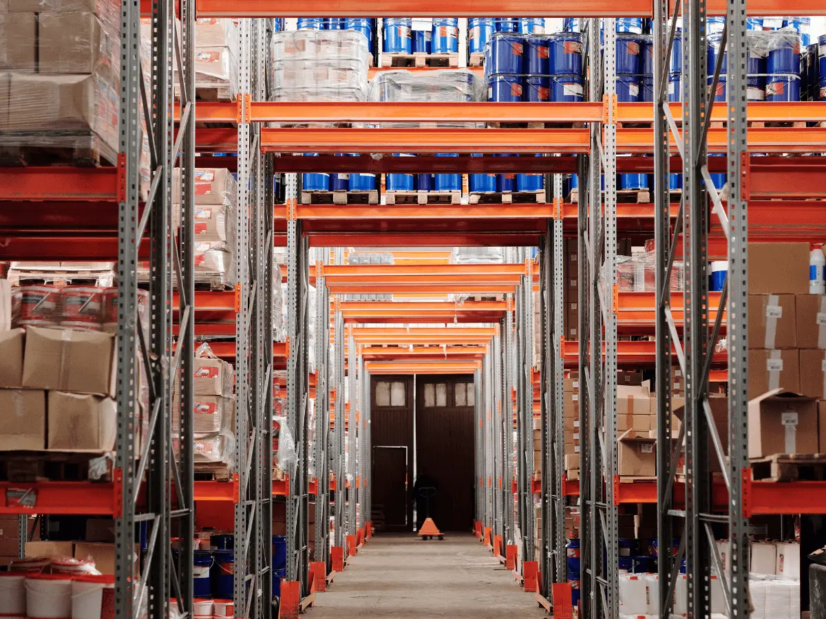 A long warehouse aisle between orange pallet racks loaded with boxes, shrink-wrapped pallets and drums, with a pallet jack at the far end